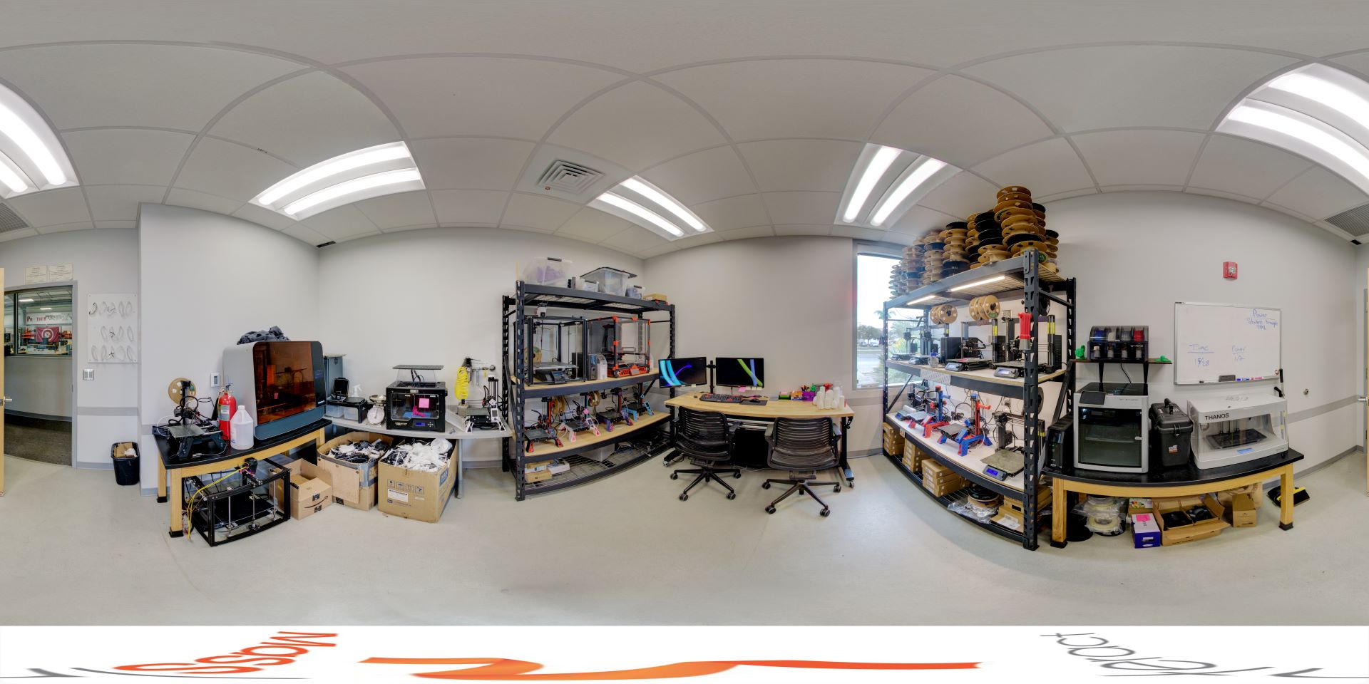 Panoramic view of a high-tech workshop featuring 3D printers, a large workbench with multiple computer monitors, and various tools and equipment organized on shelving units under bright overhead lights
